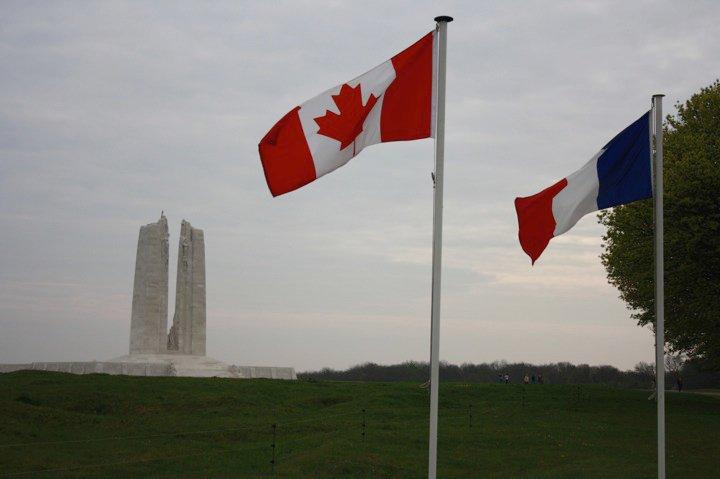 French and Canadian flags fly in the foreground, a tall white memorial monument with two large columns is in the background across a green field